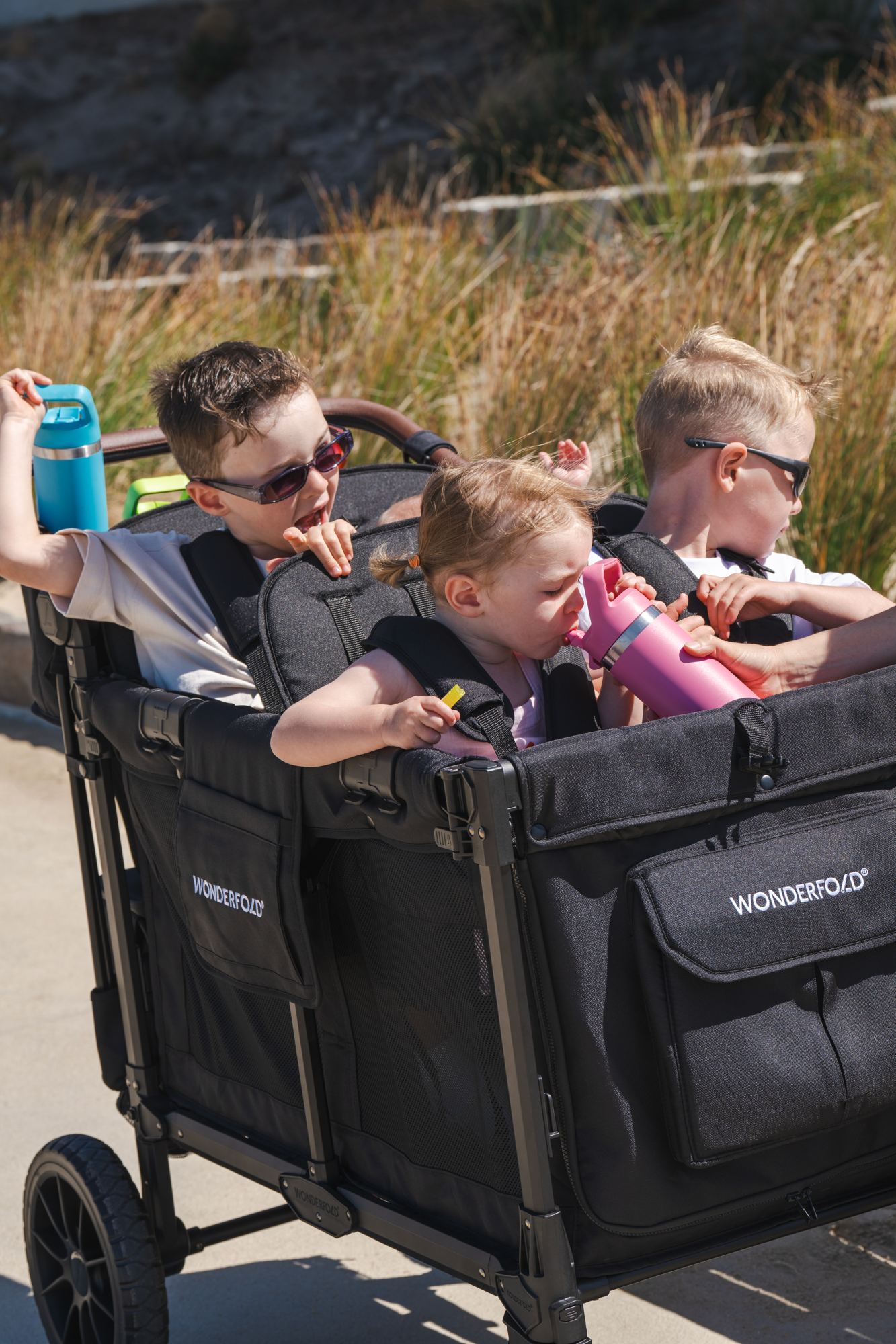 Three children with drink bottles ride in a WonderFold W4 by the beach